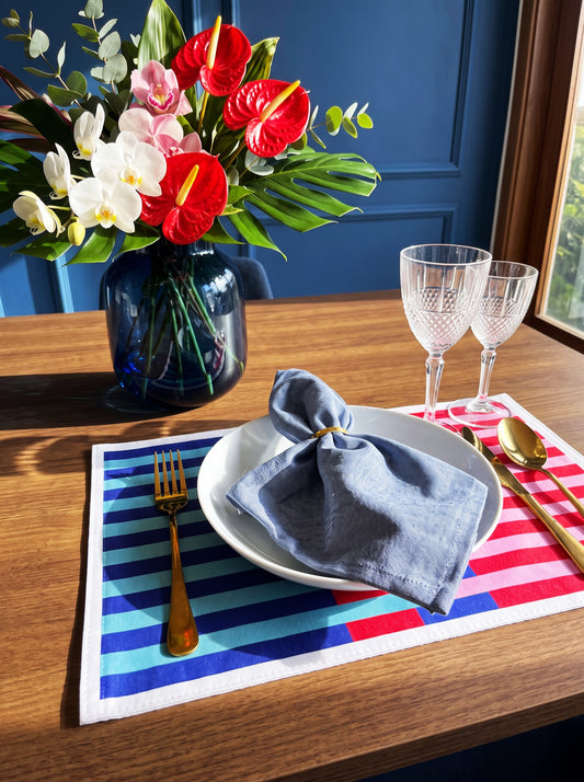 A styled dining table setting with a striped placemat in red, white, and blue, a white plate, a folded blue napkin, gold-toned cutlery, two clear wine glasses, and a dark blue vase of tropical flowers against a deep blue paneled wall.