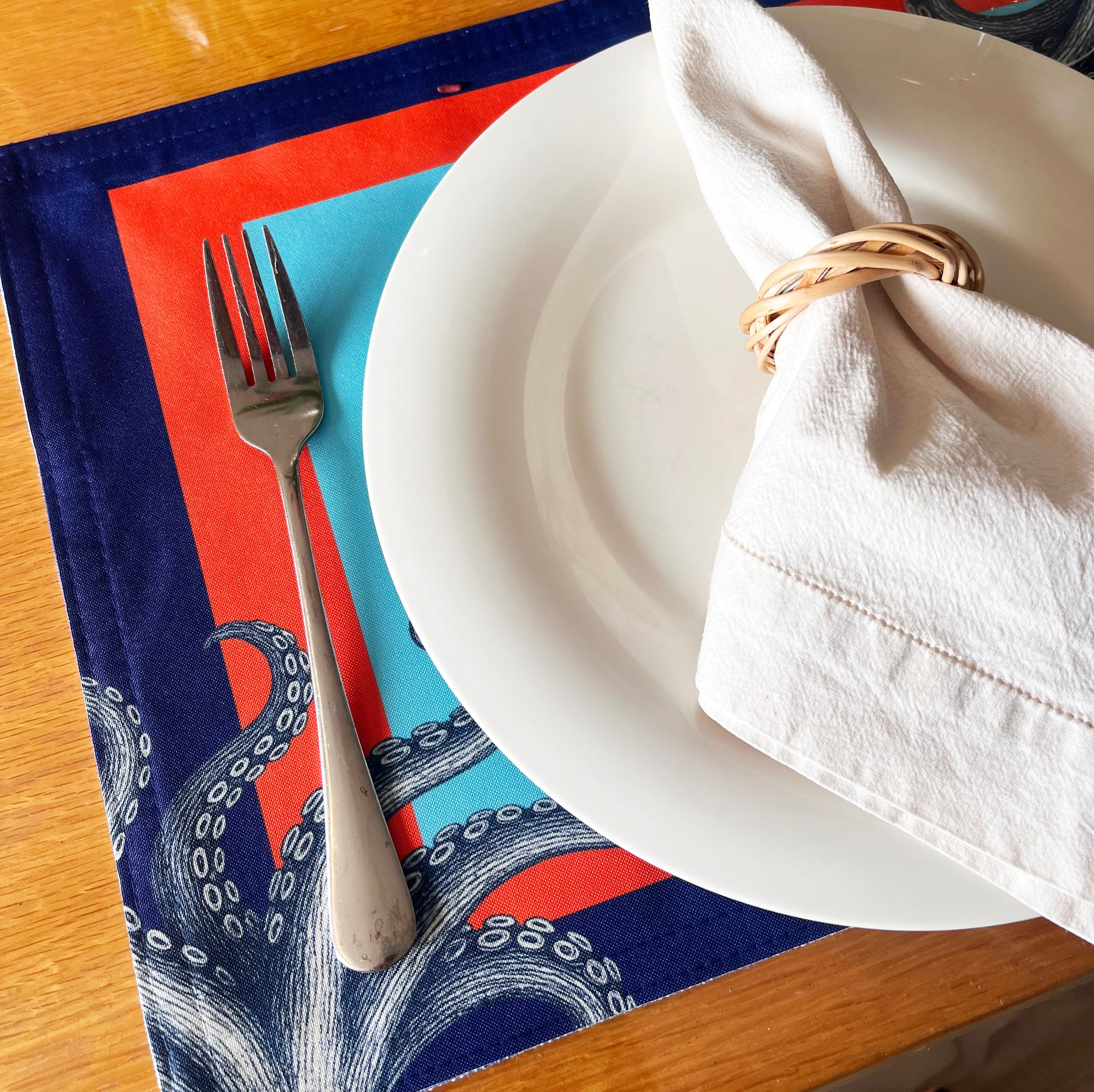 White plate with a folded white napkin, fork, and colorful placemat on a wooden table.
