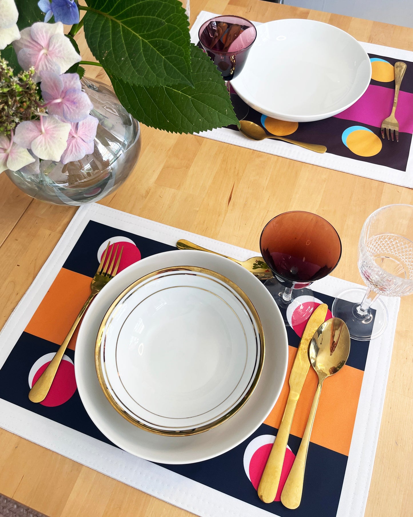 A styled dining table setting featuring a white ceramic bowl and plate set with gold trim, placed on a colorful placemat with orange, pink, and navy geometric shapes. The setting includes gold-toned flatware, a glass of red wine, a clear wine glass, and a vase of pink and white hydrangeas on a light wood table.