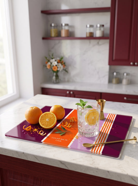 A kitchen countertop scene with a purple and orange serving board holding oranges, a glass of water with a lemon slice, a small gold jigger, a spoon, and a folded napkin. The background shows a blurred kitchen interior with dark red cabinets and a marble backsplash.