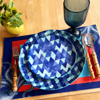 Dinner setting with blue geometric plates, cutlery, and a strong color placemat on a wooden table.