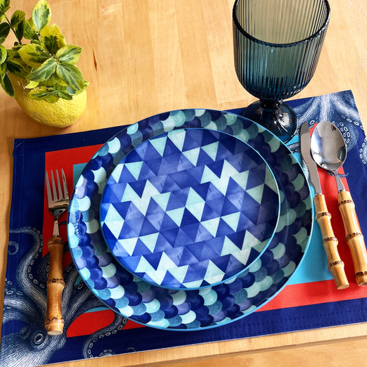 Dinner setting with blue geometric plates, cutlery, and a strong color placemat on a wooden table.