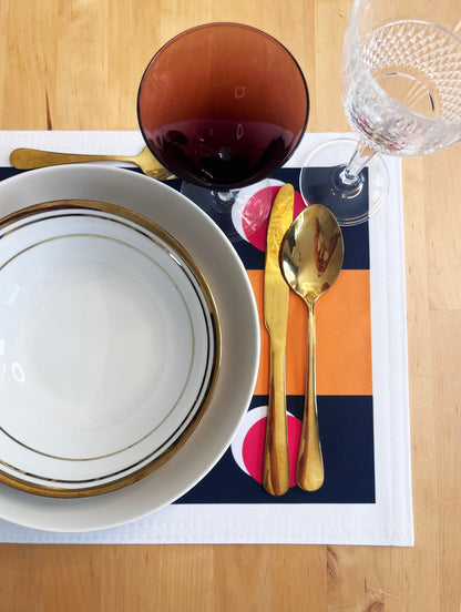 A styled table setting featuring a white ceramic bowl with gold stripes, a gold knife and spoon, a dark amber glass, and a clear wine glass on a colorful placemat over a light wood table.