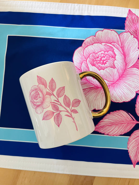 Mug with peony pink floral design on a floral-patterned placemat, golden handle. 