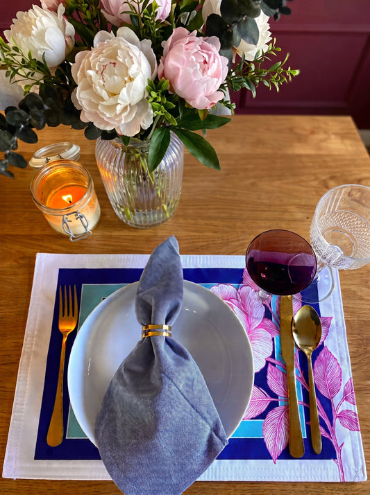 A styled tabletop setting with a white plate, folded gray napkin, gold cutlery, a pink floral placemat, a candle, a glass vase of flowers, and a glass of red wine on a wooden table.