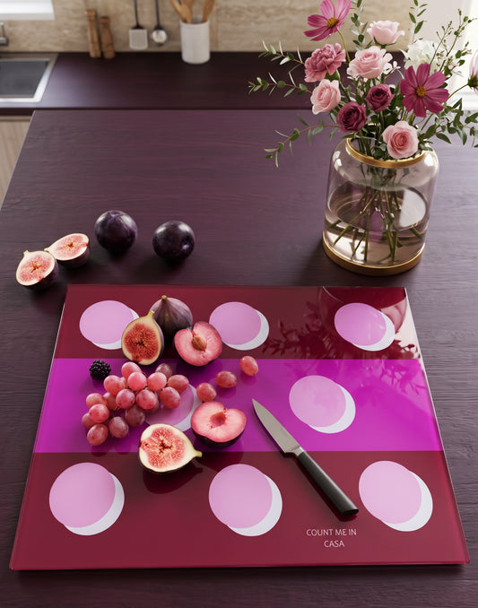 A rectangular glass cutting board with a geometric pink and magenta pattern, styled on a dark wooden kitchen countertop. The board is decorated with fresh figs, grapes, a knife, and a blackberry, with a vase of flowers and whole plums in the background.