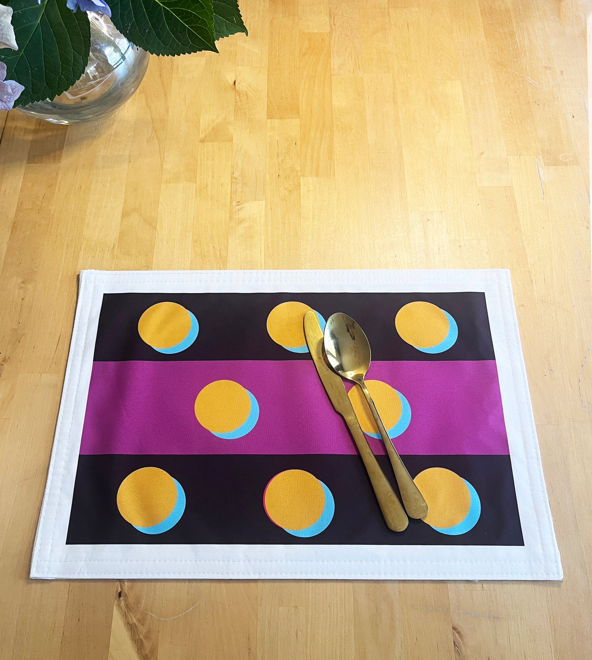 A rectangular placemat with a white border, featuring a black background with horizontal magenta stripes and yellow circles outlined in light blue. A gold knife and spoon are placed on top, set on a light wood tabletop with a glass vase visible in the top-left corner.
