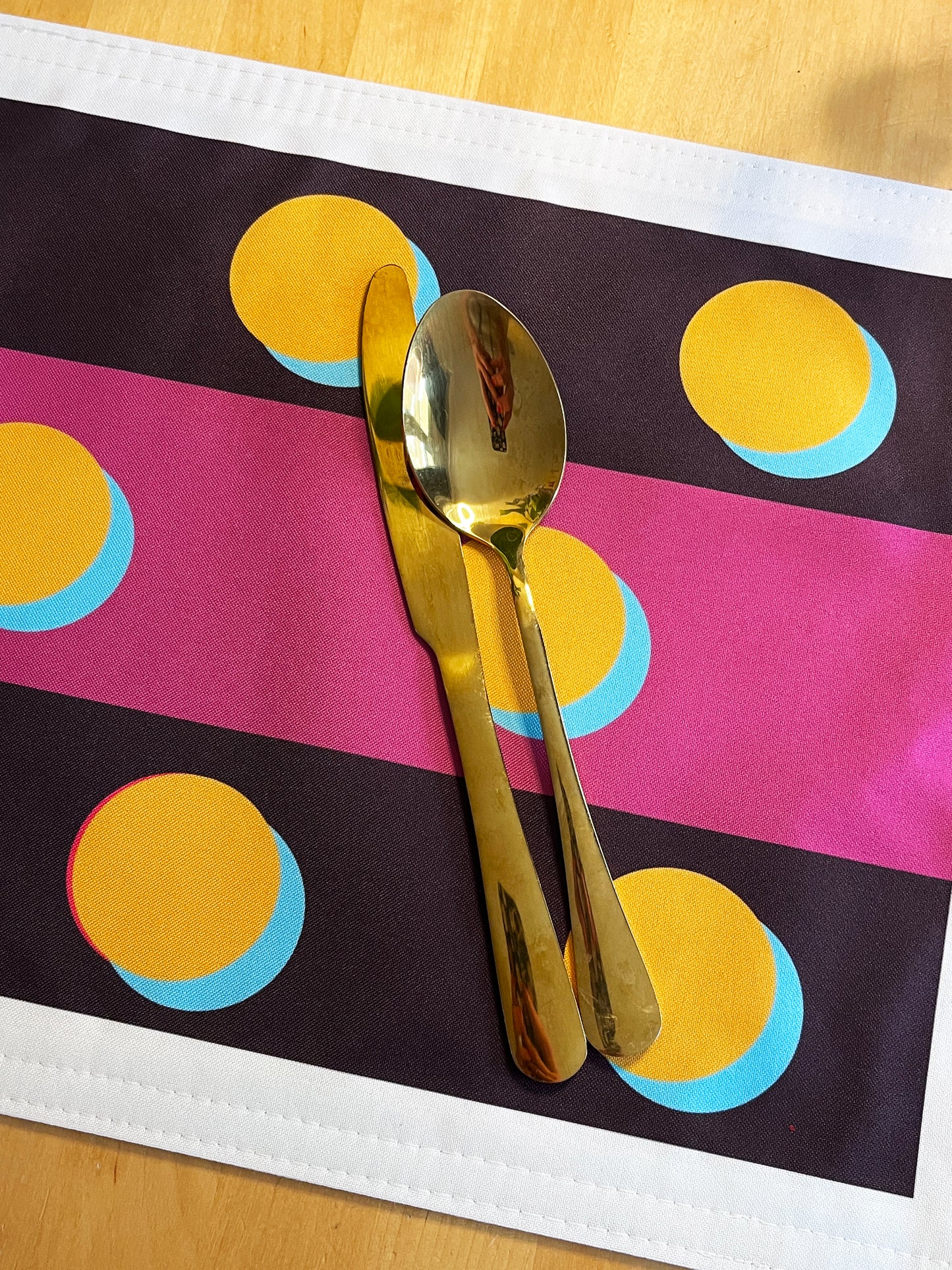 A gold metal spoon and knife set placed on a colorful placemat with pink, black, yellow, and blue stripes on a light wood surface.