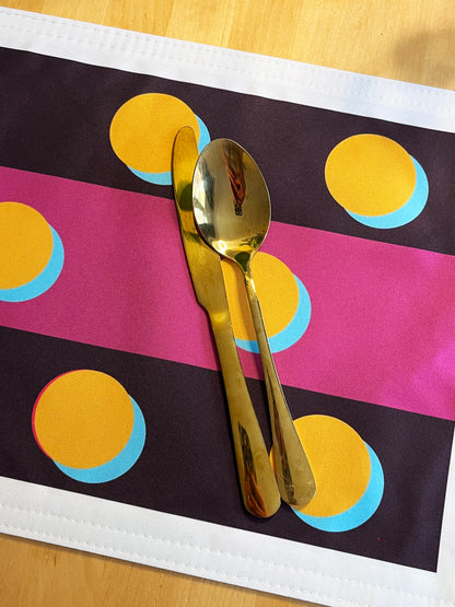 A gold metal spoon and knife set placed on a colorful placemat with pink, black, yellow, and blue stripes on a light wood surface.