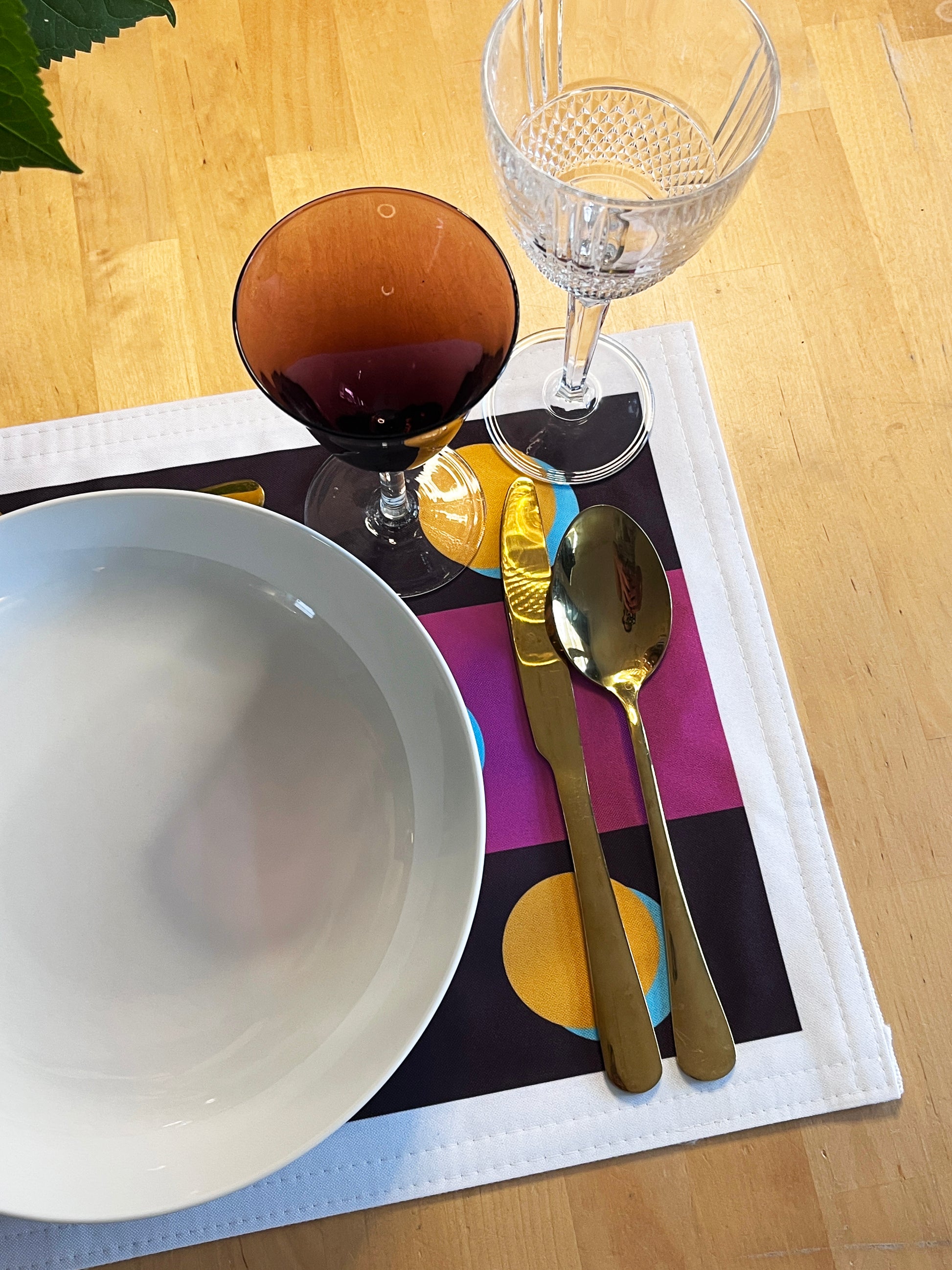 A styled table setting featuring a white ceramic bowl, gold-toned flatware (knife and spoon), a clear cut-glass wine glass, and an amber-colored wine glass, all arranged on a white placemat with a colorful geometric pattern over a light wood table.