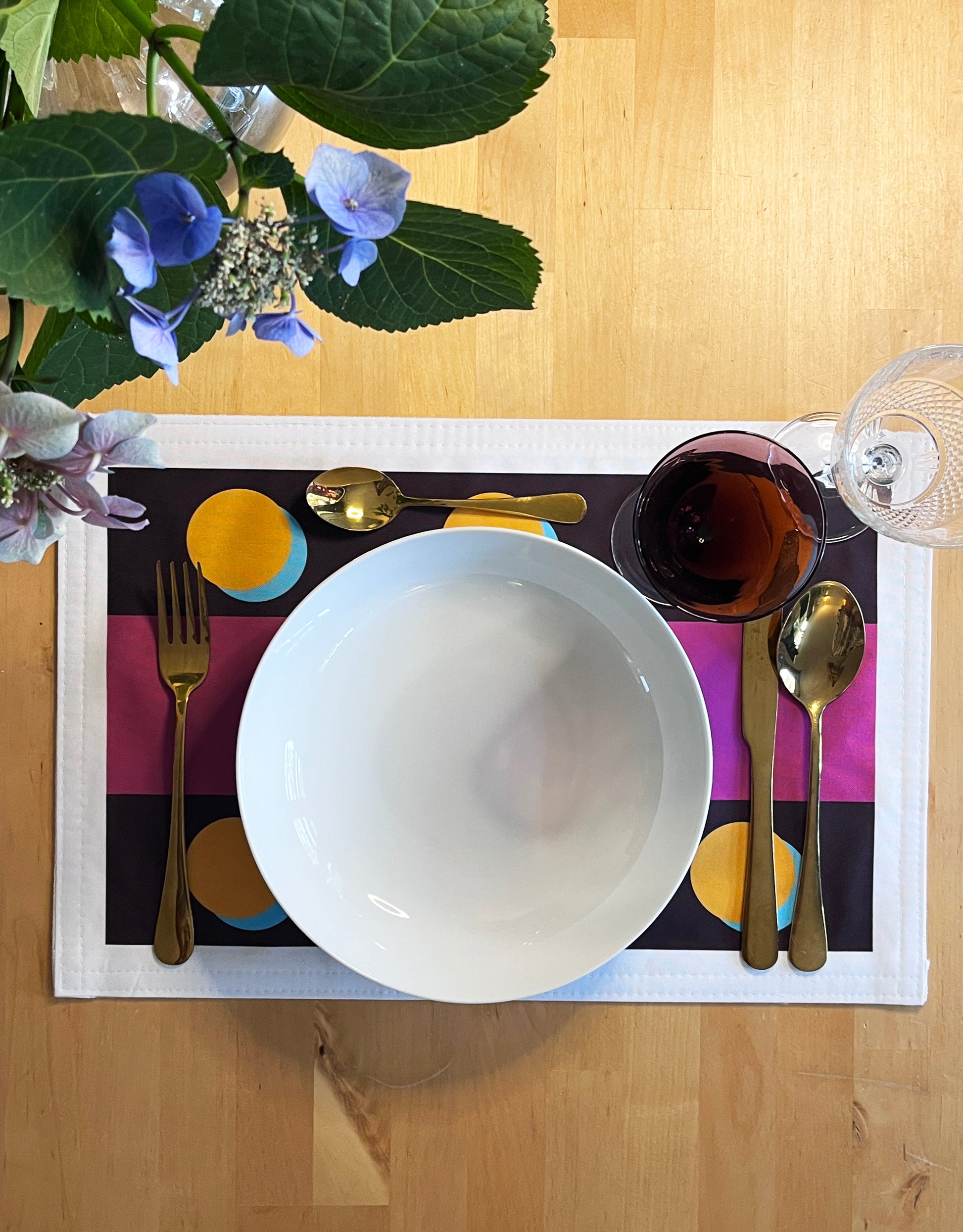 A top-down view of a place setting on a light wood table, featuring a white ceramic bowl, gold cutlery, a glass of red wine, and a colorful placemat with purple, yellow, and blue accents. A white plate and green hydrangea leaves are also visible.