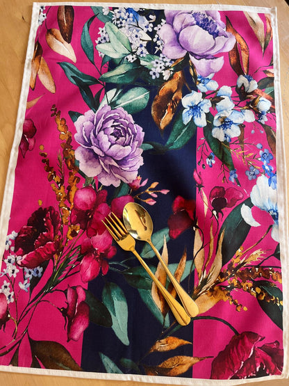 A square table linen or placemat with a vibrant floral print, styled with gold-colored cutlery on a wooden tabletop.