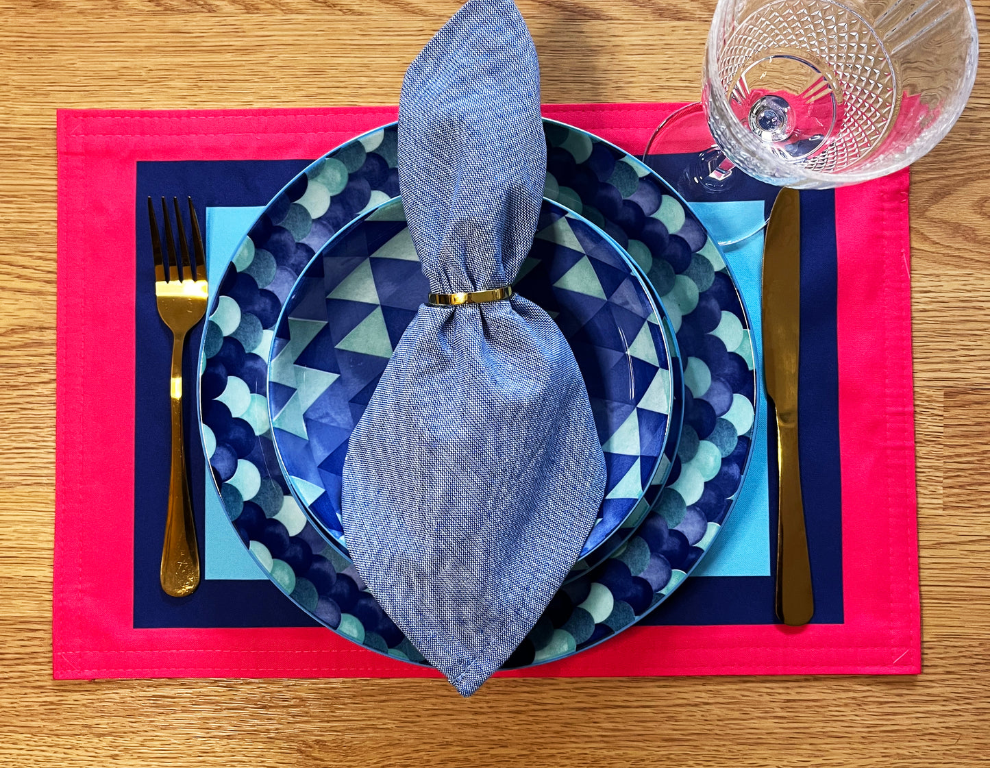 Dinner setting with blue geometric plates, silverware, and a red placemat on a wooden table with a graphic colourful placemat