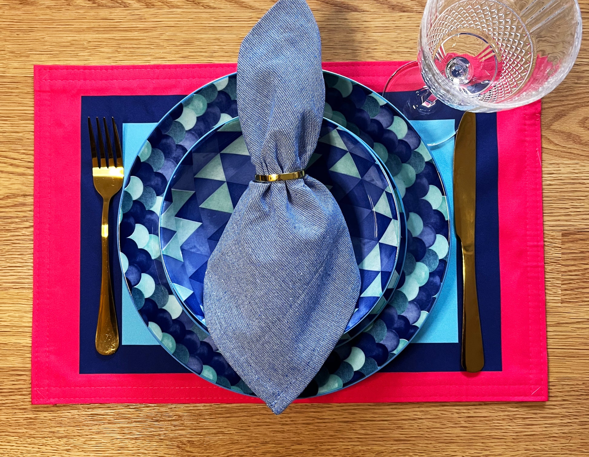 Dinner setting with blue geometric plates, silverware, and a red placemat on a wooden table with a graphic colourful placemat