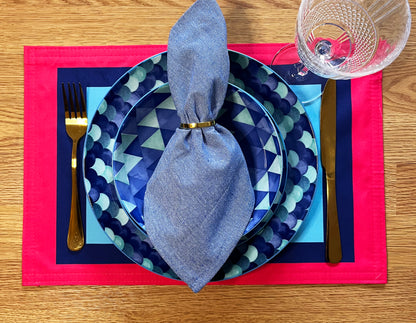 Dinner setting with blue geometric plates, silverware, and a red placemat on a wooden table with a graphic colourful placemat