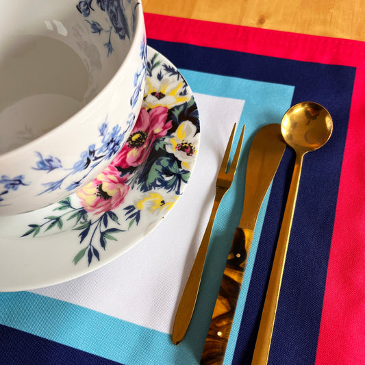 Table setting with floral plates, gold cutlery, and a colorful design in the placemat, blue, pink, and turquoise frame