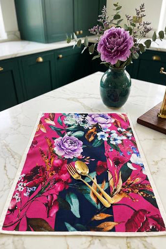 A rectangular table runner with a vibrant floral print in pink, purple, and dark blue, styled on a white marble kitchen counter with a matching green vase of flowers and gold cutlery.