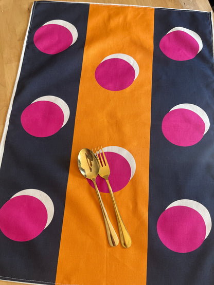 A rectangular table runner or placemat with a bold vertical stripe pattern in navy blue, mustard yellow, and magenta, featuring overlapping white semi-circles. Two gold flatware pieces are placed on the center stripe.