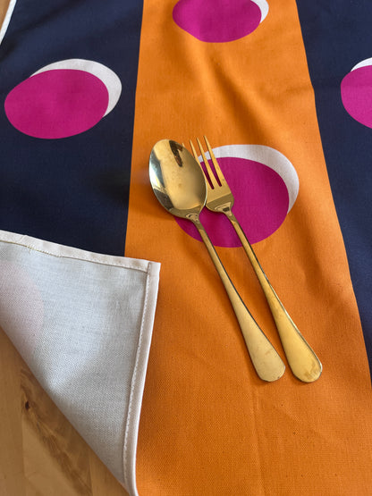 Gold-toned metal spoon and fork resting on a folded textile with a bold pattern of navy, orange, magenta, and white, with a folded-over corner revealing a light fabric underside.