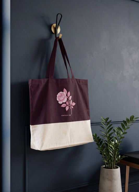 A canvas tote bag with a dark burgundy top half, cream bottom half, and a pink rose graphic, hanging from a gold hook on a dark blue wall in a styled room setting.