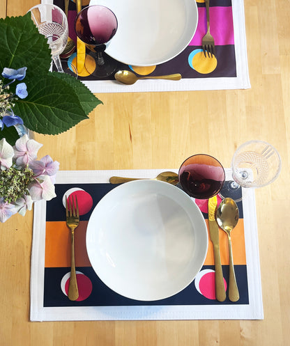 A top-down view of a table setting featuring two matching placemats with a bold geometric pattern of orange, pink, yellow, and dark blue blocks. Each placemat is styled with a white bowl, gold cutlery, a glass of red wine, and a glass, set on a light wood table next to green and purple flowers.