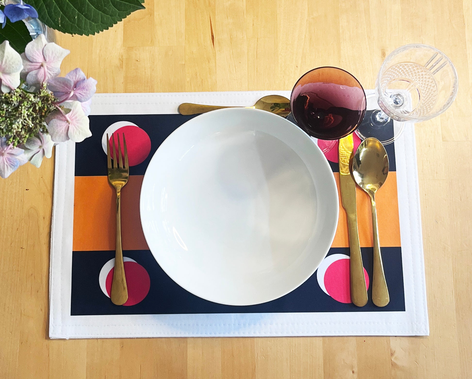 A styled table setting featuring a rectangular placemat with a black, orange, and pink geometric pattern, a large white ceramic plate, gold-toned cutlery, a glass of red wine, and a clear drinking glass on a light wood table.
