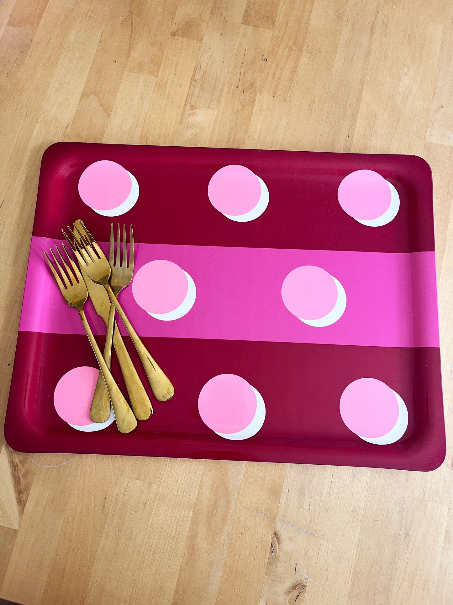 A rectangular tray with rounded corners, featuring a dark red and bright pink striped pattern with pink polka dots, styled with four gold forks on a light wood floor.