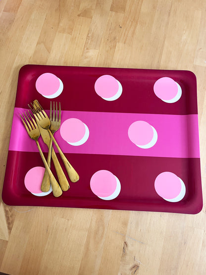 A rectangular tray with rounded corners, featuring a dark red and bright pink striped pattern with pink polka dots, styled with four gold forks on a light wood floor.