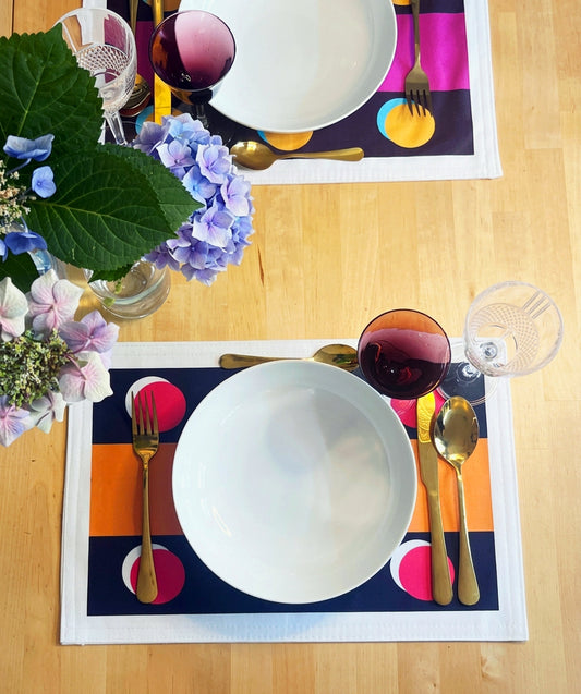 Two place settings on a light wood table, featuring colorful striped placemats with white bowls, gold cutlery, and wine glasses, alongside a floral centerpiece.