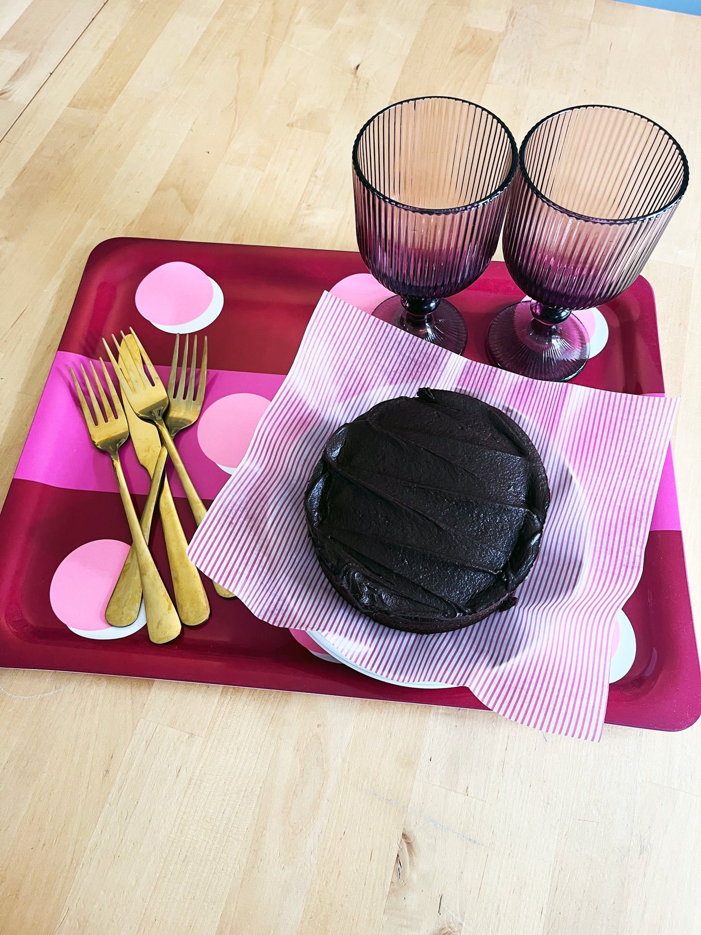 A round chocolate cake with thick frosting sits on a pink-and-white striped paper napkin on a red tray, alongside two gold-colored forks and two ribbed purple glass goblets, all arranged on a light wood tabletop.