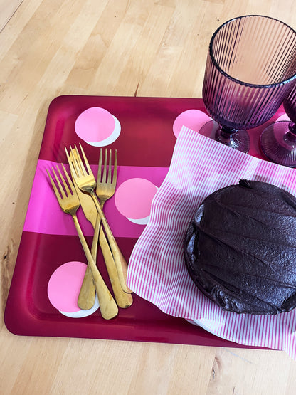 A styled tabletop scene on a light wood surface showing a dark chocolate cake on pink-and-white striped paper, three gold forks on a magenta tray, and two purple ribbed drinking glasses.