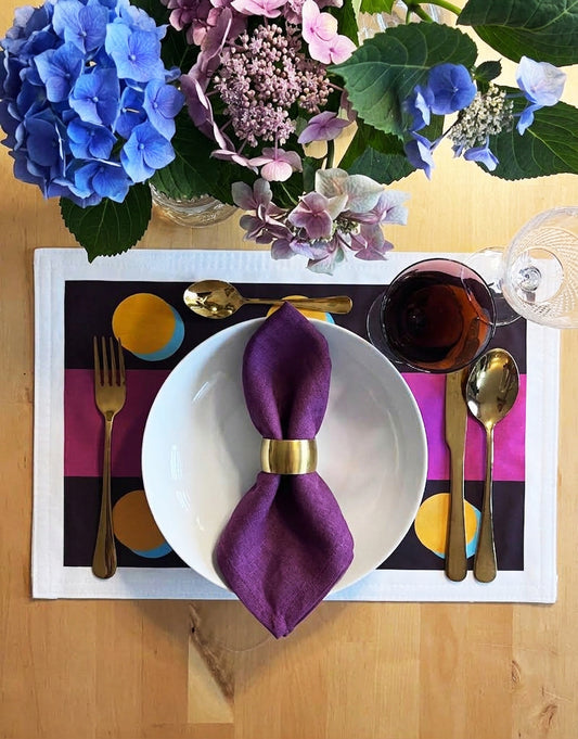 A styled tabletop place setting on a light wood surface, featuring a white plate with a purple napkin tied with a gold ring, gold cutlery, a glass of red wine, and a colorful placemat. A floral arrangement with blue and pink hydrangeas sits above the setting.