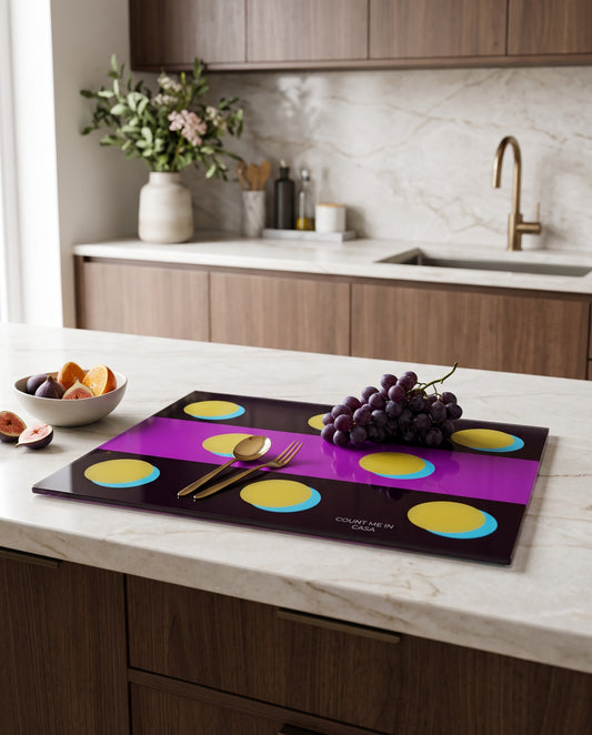 A rectangular glass serving platter with a purple center band and yellow and blue circular patterns, placed on a marble kitchen island. The platter holds gold-toned cutlery and a bunch of purple grapes, with a bowl of fruit and a modern kitchen in the background.