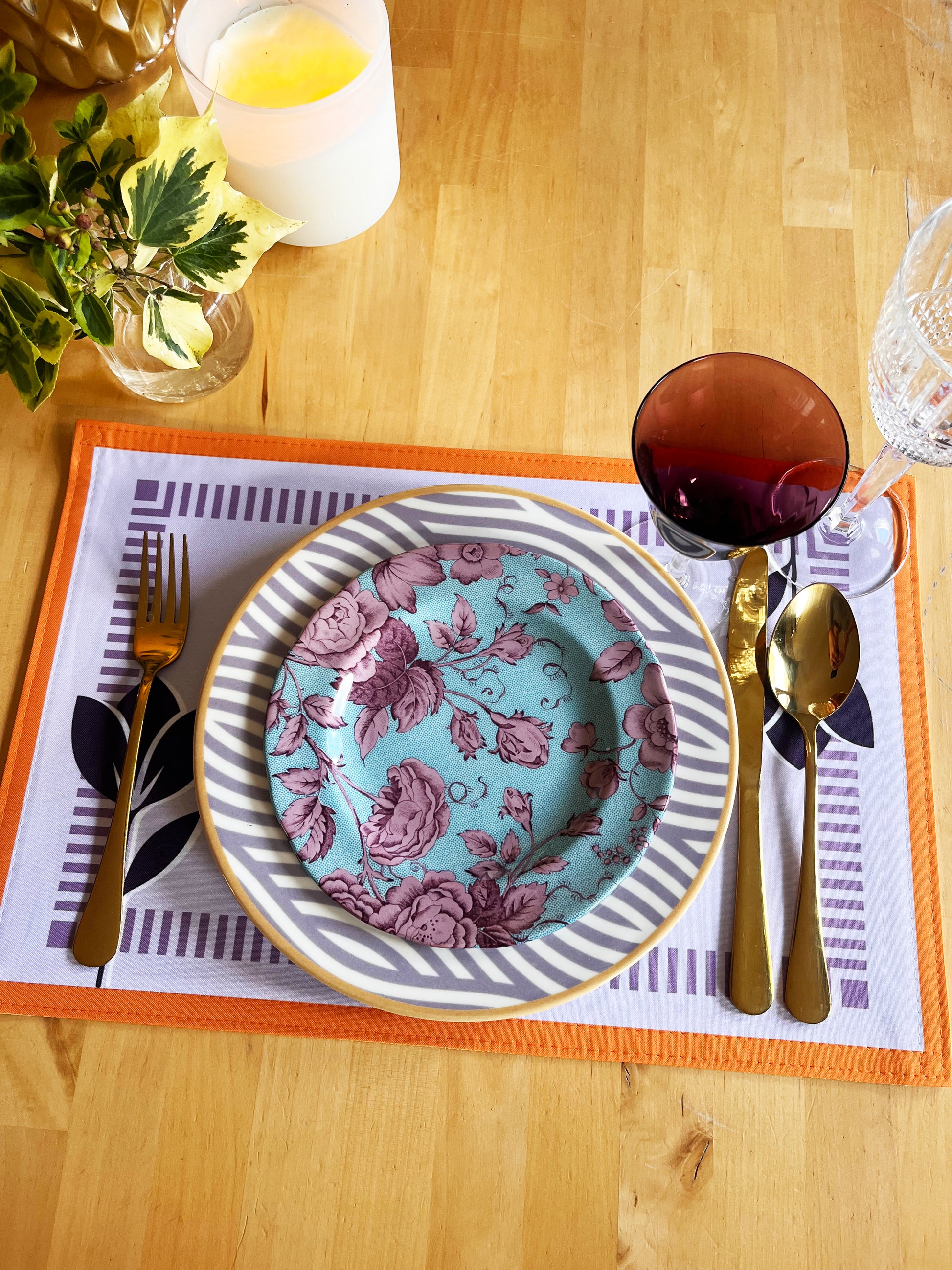 Dinner setting with floral plate, cutlery, and placemat on a wooden table. Purple flowers and orange frame. Exclusive design