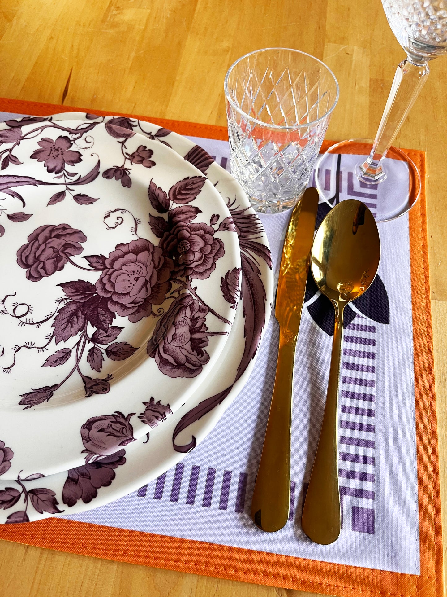 Table setting with floral plate, gold cutlery, and clear glass on an orange placemat, zoom