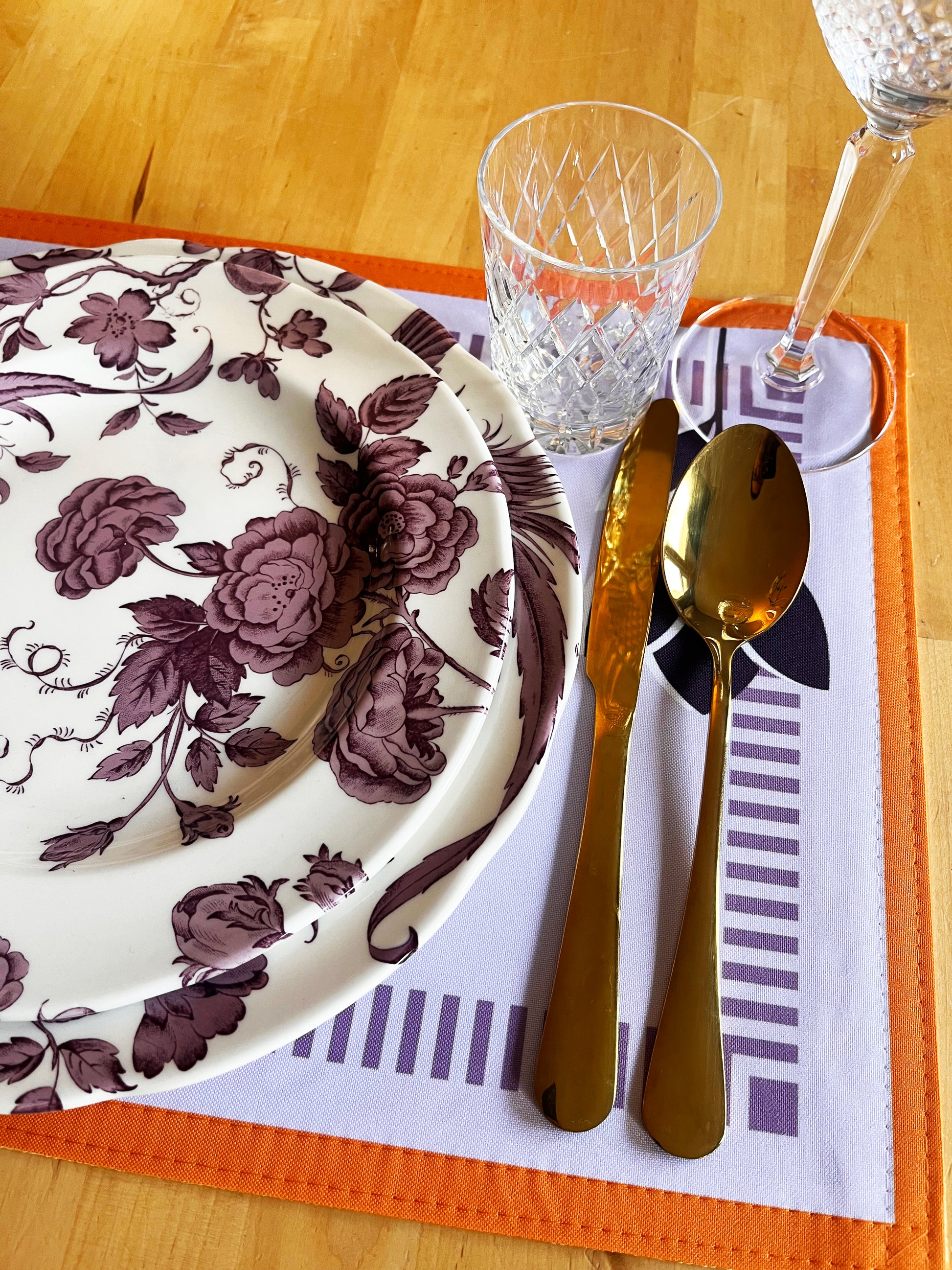 Table setting with floral plate, gold cutlery, and clear glass on an orange placemat, zoom