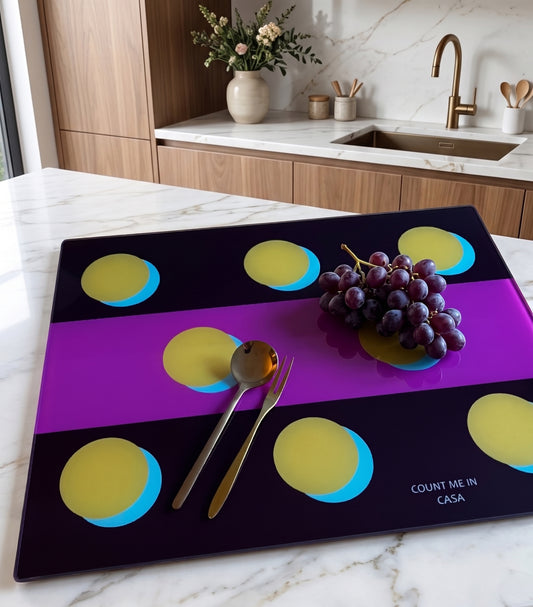 A rectangular glass serving platter with a modern geometric design of purple and black horizontal bands, yellow circles, and blue gradients, placed on a white marble kitchen countertop. The platter is styled with silver cutlery and a bunch of purple grapes, with a kitchen background.