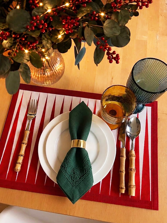 A festive tabletop setting featuring a red and white striped placemat with a white plate, a dark green textured napkin tied with a gold ring, and bamboo-handled cutlery. A matching red placemat is also visible on the table, alongside a green ribbed tumbler, an amber glass, and a holiday wreath in the background.