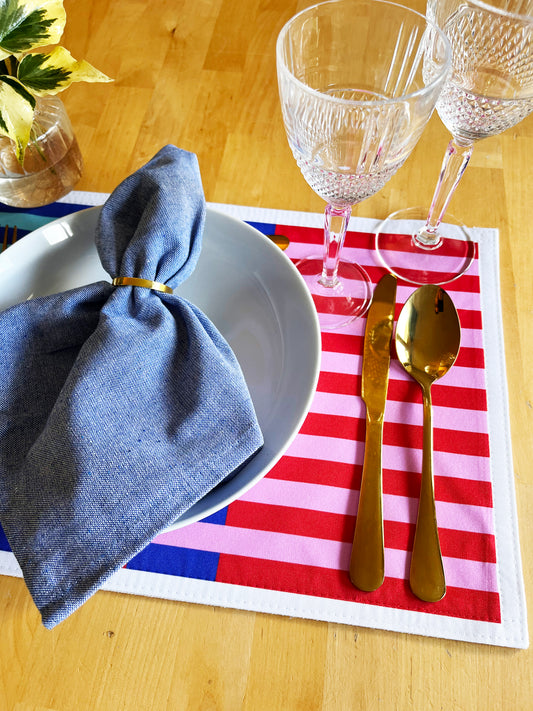 Dinner setting with blue napkin, white plate, gold cutlery, and red and white striped placemat on a wooden table.A stylish, versatile collection designed for modern table settings, home décor lovers, and anyone who enjoys effortless creativity.
