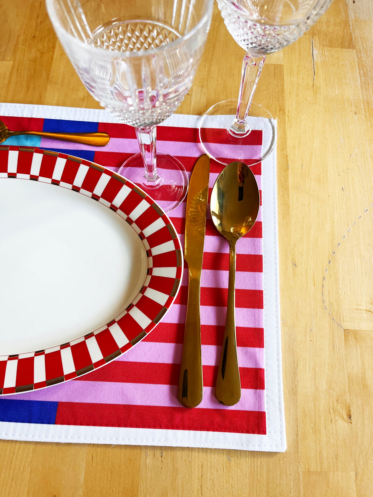 Dinner setting with a blue, red, and pink striped placemat, wine glasses, knife, and spoon on a wooden table. Play with beautiful combinations, express your personal touch, and bring more colour, joy, and personality to your table.