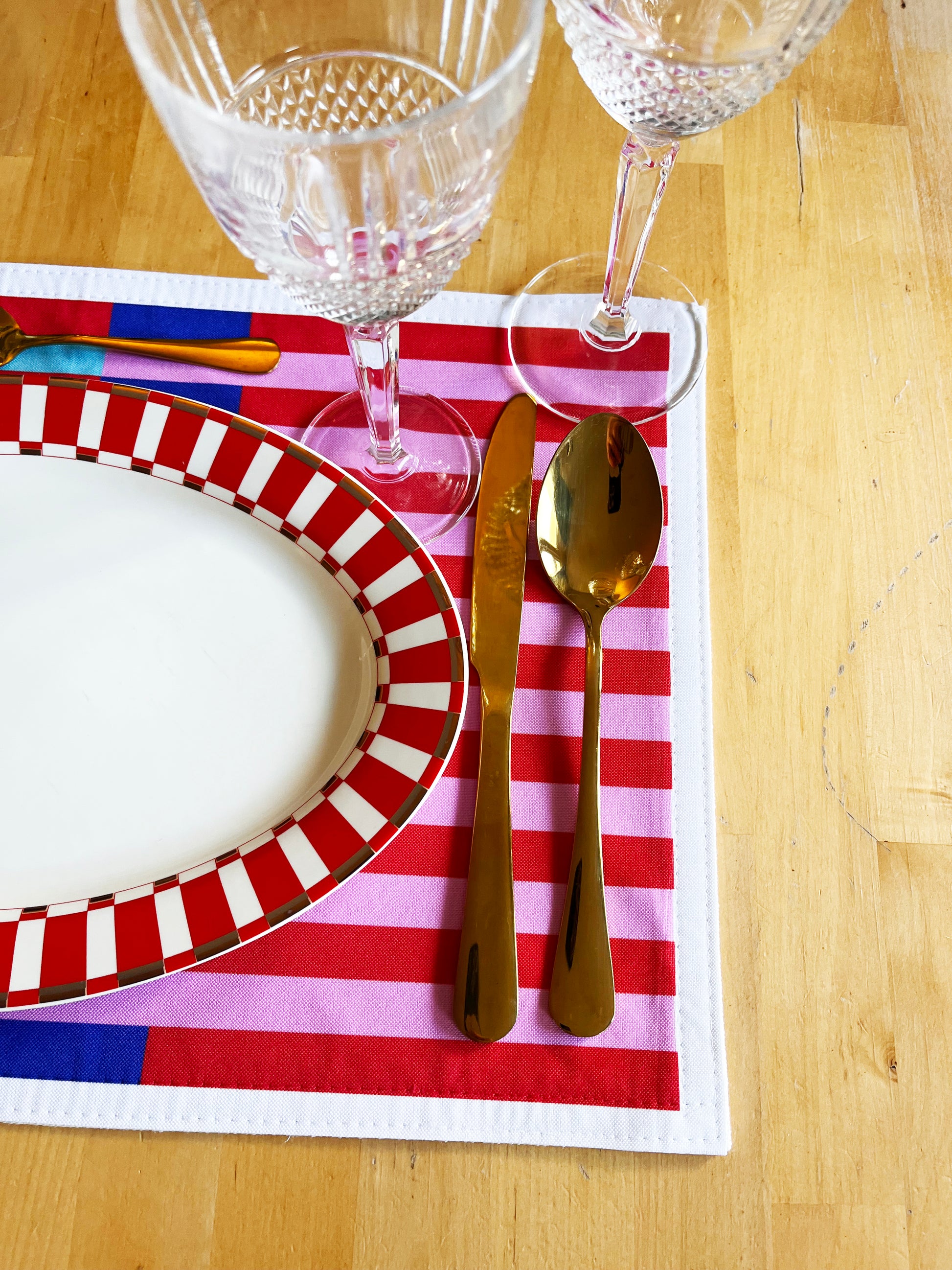 Dinner setting with a blue, red, and pink striped placemat, wine glasses, knife, and spoon on a wooden table. Play with beautiful combinations, express your personal touch, and bring more colour, joy, and personality to your table.