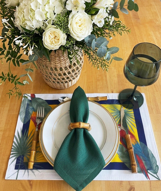 A styled table setting featuring a tropical print placemat with a white plate, a green napkin tied with a bamboo ring, cutlery, and a dark green wine glass on a wooden table. A woven basket vase filled with white flowers and greenery sits in the background.