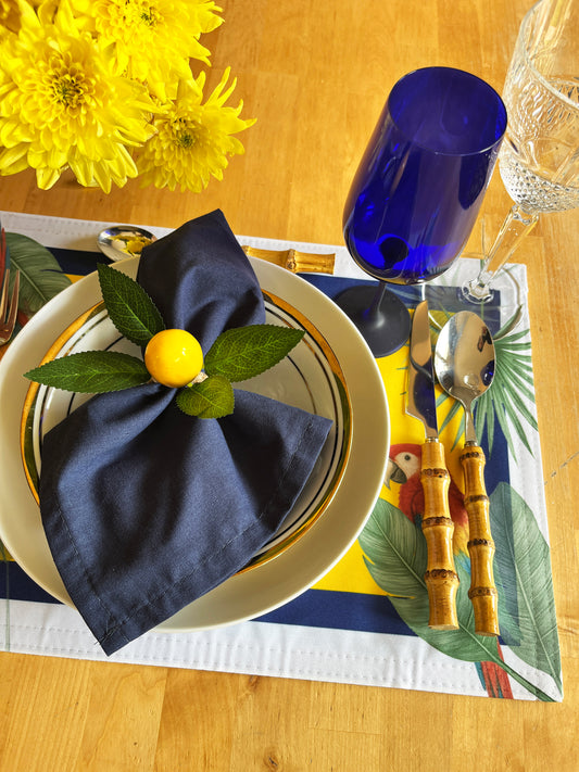 A styled tabletop setting on a wooden table featuring a navy blue napkin with a small yellow pom-pom and green leaves, a white plate with gold trim, bamboo-handled silver cutlery, a blue tumbler, and a tropical placemat.