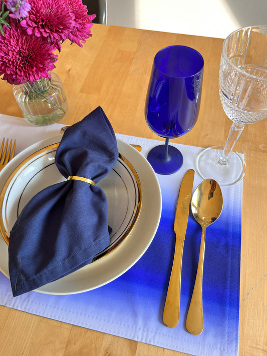 A styled table setting on a light wood table featuring a blue and white gradient placemat, a navy blue napkin tied with a gold band, white plates with gold trim, gold flatware, a cobalt blue wine glass, and a clear wine glass. A small vase of pink flowers is visible in the background.
