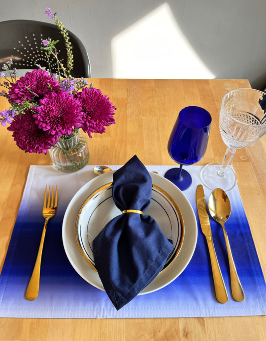 A styled dining table setting with a blue and white gradient placemat, a white plate with gold trim, a navy blue napkin, gold flatware, a cobalt blue wine glass, and a clear wine glass, alongside a vase of magenta flowers on a wooden table.