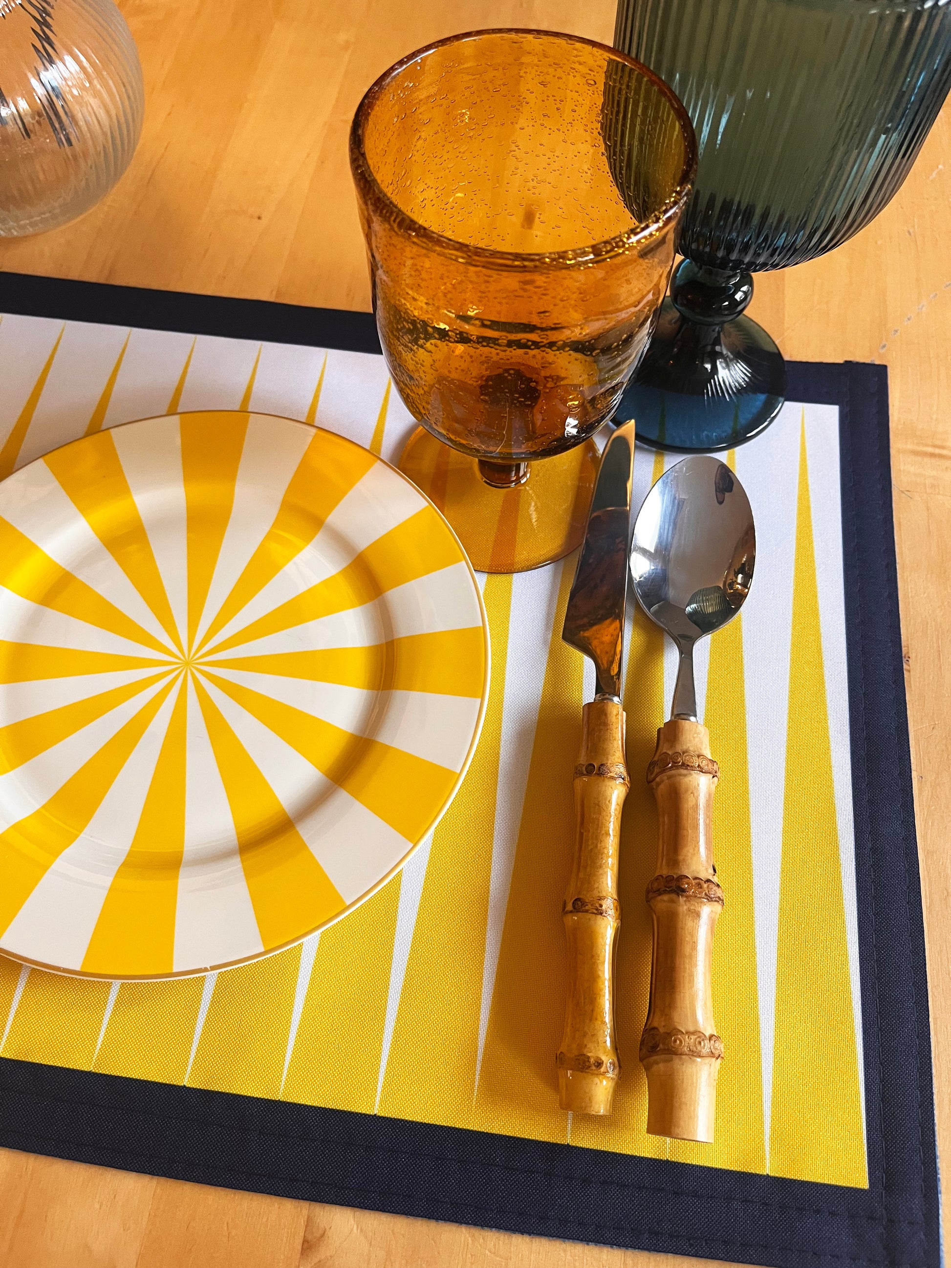 Yellow and white plate with a sunburst pattern on a yellow placemat, accompanied by a brown glass, knife, and spoon.