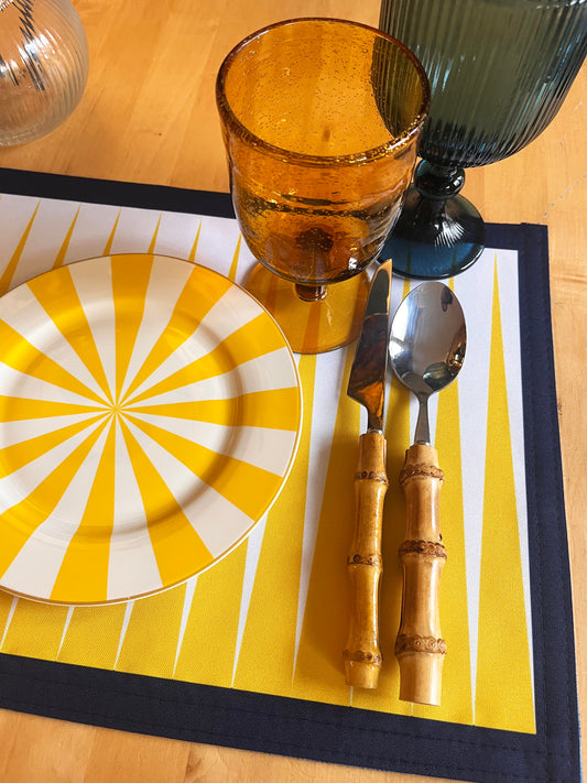 Yellow and white plate with a sunburst pattern on a yellow placemat, accompanied by a brown glass, knife, and spoon.