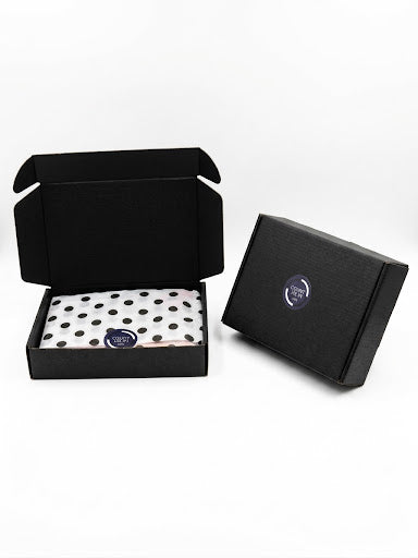 Two black branded gift boxes on a white background, with one open box showing a white tissue or paper insert with black polka dots and a small round blue logo, and the other closed box showing a matching blue logo on the lid.