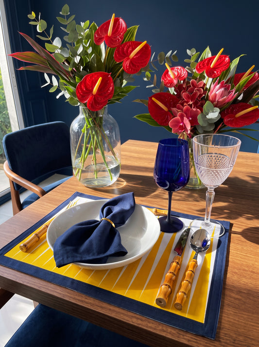 A styled table setting featuring a yellow and white striped placemat with a navy blue border, a white bowl with a navy blue napkin, bamboo handled cutlery, a cobalt blue tumbler, a clear wine glass, and a vase of red flowers on a wooden table.