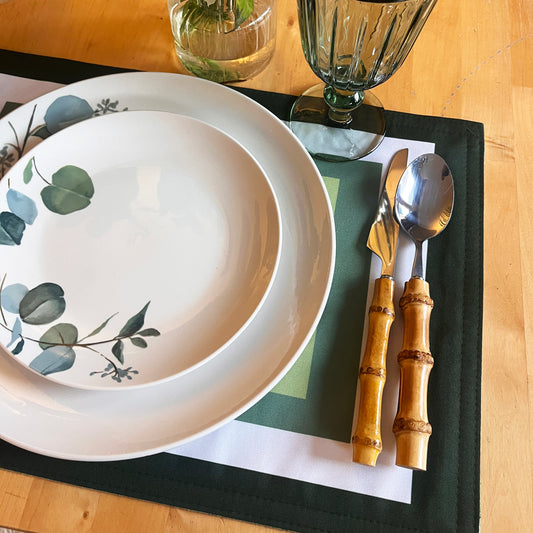 Dinner setting with a floral-patterned plate, bamboo cutlery, and green glass goblet on a wooden table, shades of green frame placemat.